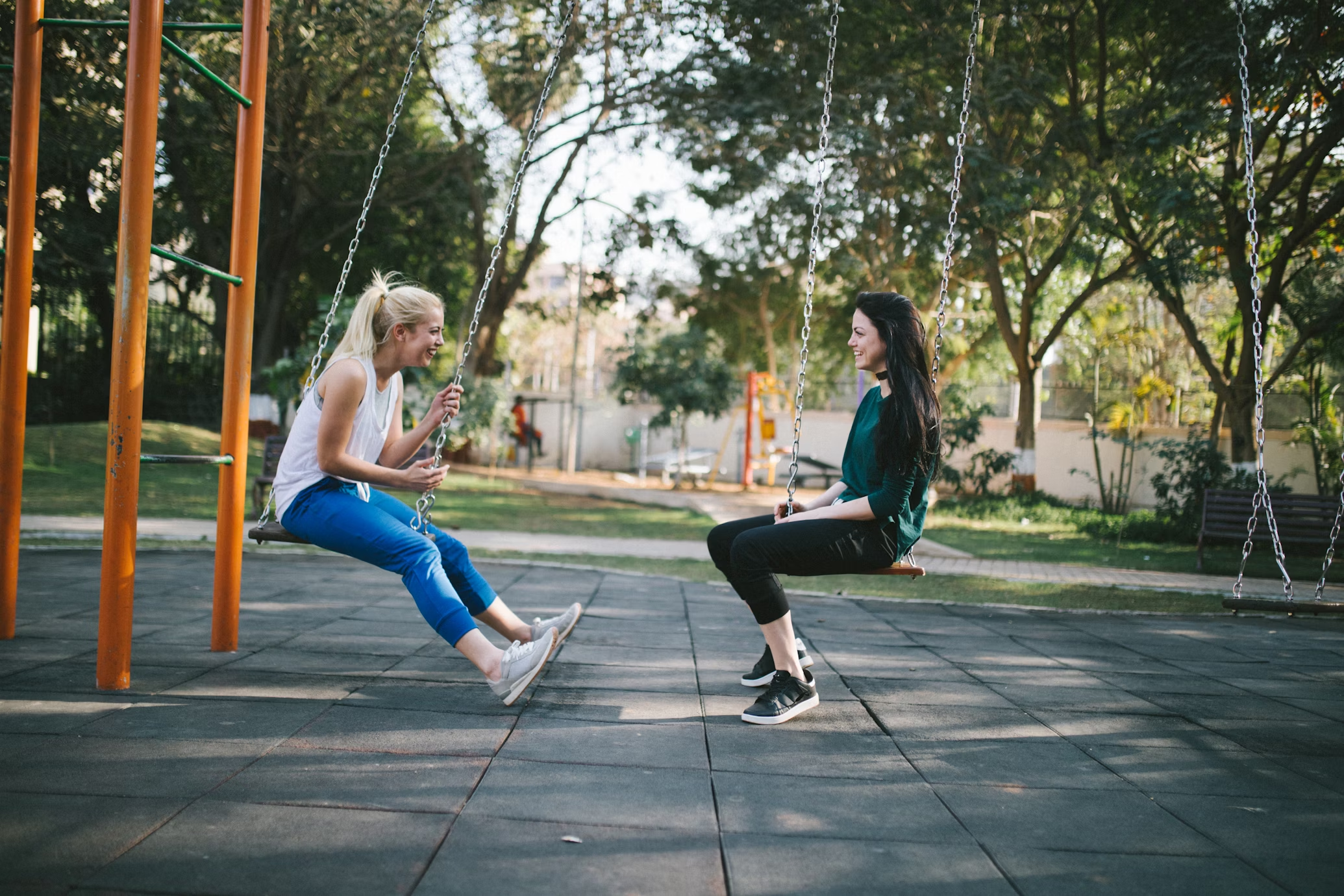 Two girls playing on a swing set in a park surrounded by lots of stainless steel equipment.